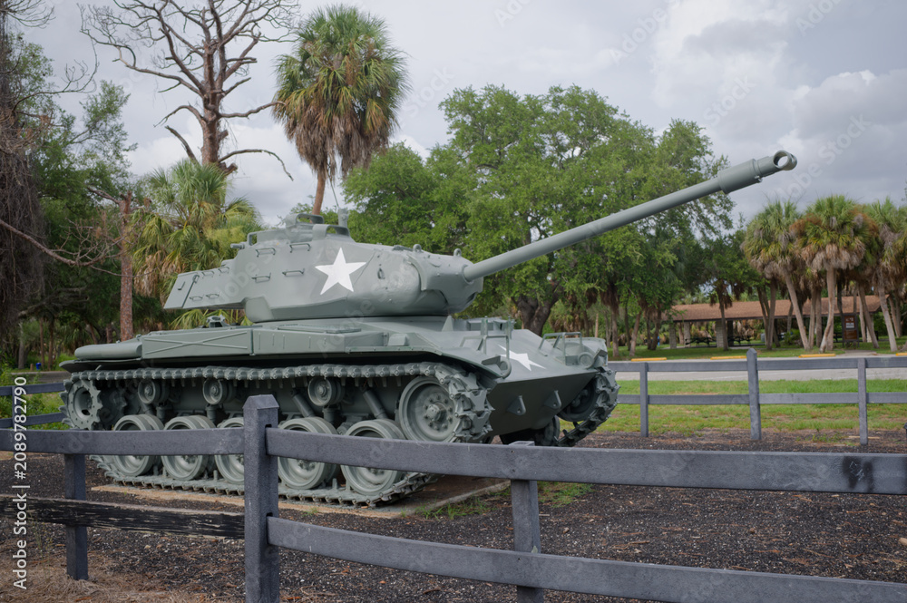 Fototapeta premium A restored historic military tank monument sits on display in a park setting with palm trees and wooden fencing. War Veterans Memorial Park In St. Petersburg, FL conveying military history, remembranc