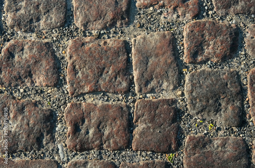 Top down view of irregular square brownish-red granite cobblestone pavement on a city sidewalk in bright sunlight, with fine gravel in the joints