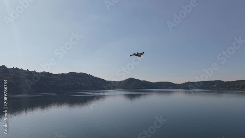 Drone aerial view of a firefighting aircraft flying in the distance over a mountain and lake landscape under a clear blue sky. Scenic telephoto-style footage suitable for aviation, emergency response,