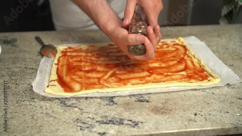 Man preparing homemade pizza, adding spices over tomato sauce on dough.