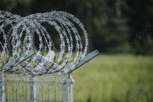 Slika na platnu Barbed wire fence coils atop a metal structure with a blurred green field in the