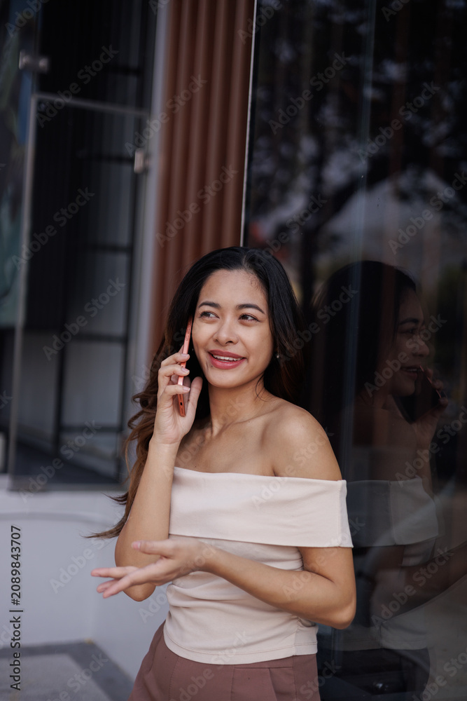 Fototapeta premium Young Asian Woman Smiling While Talking on Smartphone Outdoors Near Modern Building