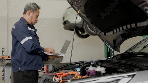 Asian senior mature EV car engineer male worker service monitor DC Inverter check with laptop computer in EV Garage auto service