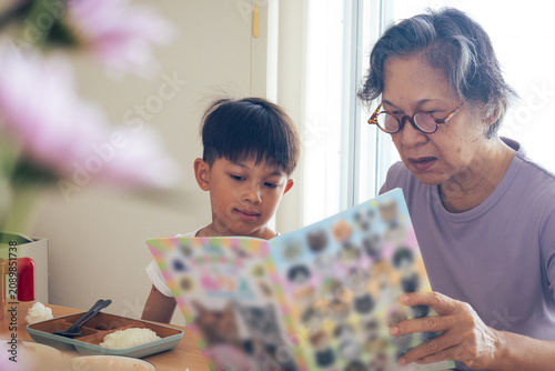 Fotografi An Asian grandmother wearing eyeglasses reading an illustrated book with her cute young grandson at the table, sharing an educational and heartwarming family bonding moment inside a bright room