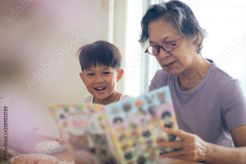 Konstfotografi An Asian grandmother wearing eyeglasses reading an illustrated book with her cute young grandson at the table, sharing an educational and heartwarming family bonding moment inside a bright room