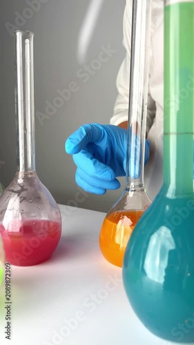 A person wearing blue gloves pours orange liquid into a clear container with red liquid. Green and blue liquids are also present in various flasks.