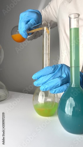A person in gloves pours orange liquid into a glass container. The colors change and mix in various containers. The activity shows chemical reactions.