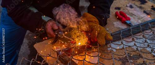 Schilderij op canvas A worker wearing protective gloves and a jacket welds metal wire mesh outdoors; bright sparks and smoke rise as the joints are welded during repairs and maintenance