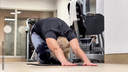 Woman with short blonde hair practices yoga on a mat in a gym, moving from a forward bend to a tabletop position with fitness equipment visible