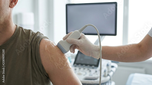 Ultrasound scan on shoulder with gel applied, clinician hands demonstrating medical diagnostic procedure in clinic room with equipment