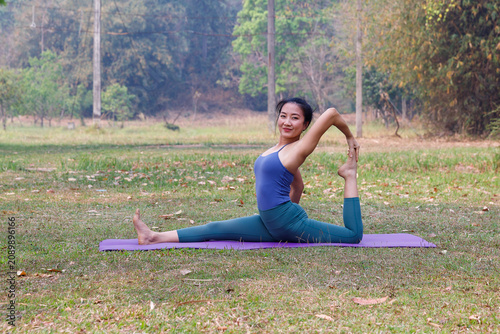 Smiling young Asian woman practicing mermaid yoga pose (king pigeon posture) on purple mat on green grass in a peaceful park.