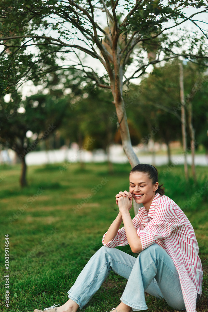 Fototapeta premium Cheerful woman outdoors, sitting on grass, smiling softly, relaxed posture, casual striped shirt, jeans, sunlit moment, gentle breeze, natural colors, serene park background