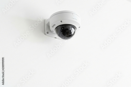 Security Dome Camera Mounted on Wall: A modern, white dome security camera is securely mounted on a plain white wall, its dark lens aimed outward, suggesting surveillance and safety.