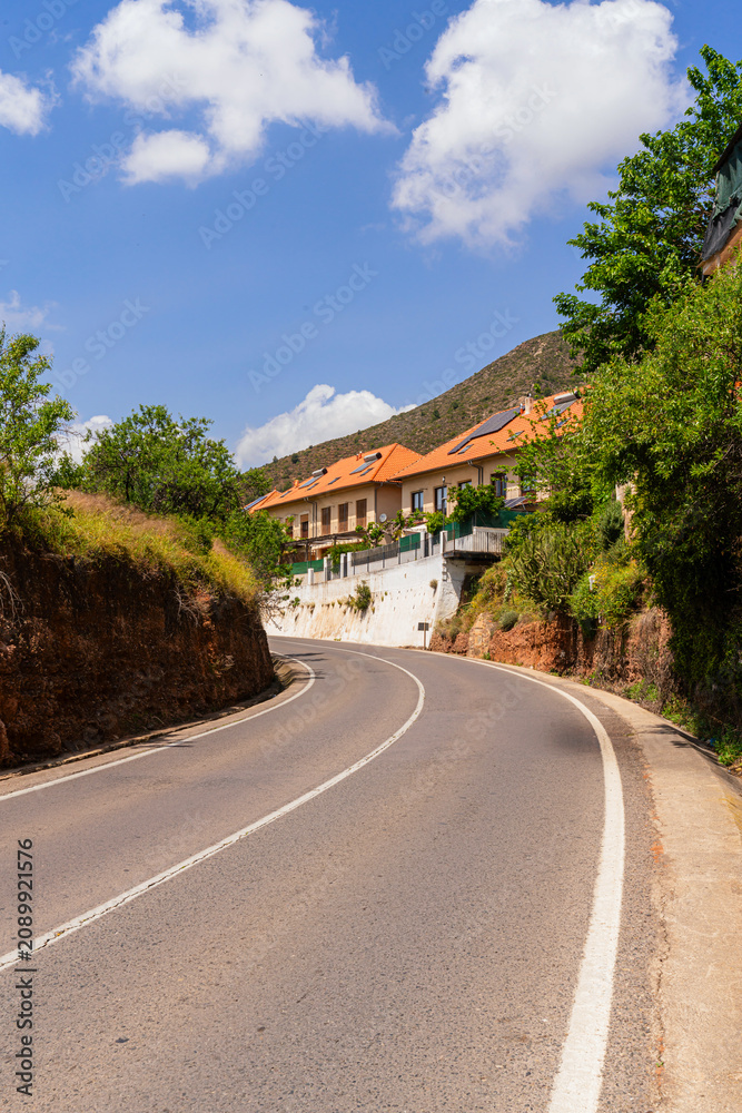 Naklejka premium Sundrenched curve of a quiet rural road leads through a hillside village, with terracottaroofed homes, lush greenery, and mountain backdrop evoking authentic slow travel in Mediterranean.