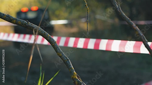 Selective focus on tree branch with red and white barrier tape in background. Blurred off-road vehicle with glowing tail lights on dirt track. Scene depicts mud racing competition in nature