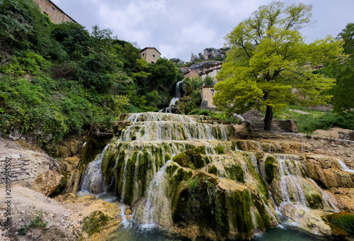 Waterfall flowing down, in rural landscape with stone buildings and greenery, with waterfall, stone steps, trees, buildings, with rock, moss, wood texture, with green, brown accents.