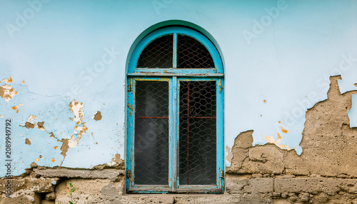 Close-up shot of a weathered wall with an arched window recessed into the wall, dark window frame, vibrant turquoise blue paint, heavily peeled paint, extensive cracks and distressed surface texture