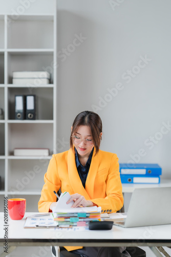 Focused Professional at Work: A woman in a vibrant orange blazer meticulously reviews documents at her desk, surrounded by office supplies, including a laptop and calculator, in a bright.
