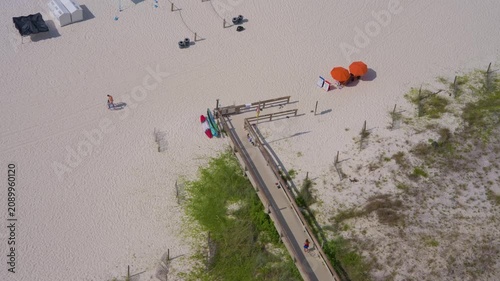 Aerial view of a wooden beach boardwalk leading to pristine white sand in Gulf Shores / Orange Beach, Alabama. People walk along the path toward bright orange umbrellas and the Gulf of Mexico beach.