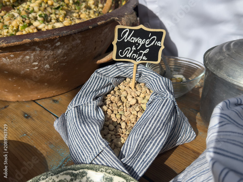 A striped fabric bag of raw manarolia or mpiza legumes with a Greek chalkboard sign, an ingredient for traditional fotokolyva salad on a rustic table.