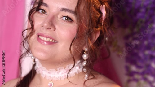 Video close up. 60 fps. Mature white woman with bare shoulder, choker on neck, pearl earrings, pink dress, hand fan smiles, looks at camera near lilac wisteria flowers. Fashion, diverse beauty, magic