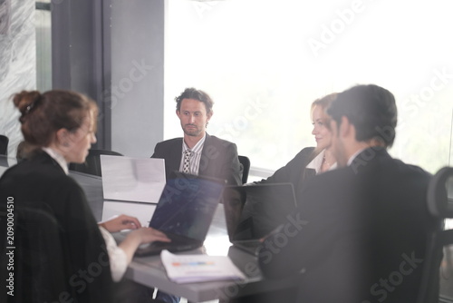 Foto Ginger-haired businesswoman types on a laptop at a sunlit boardroom table while a bearded colleague and other teammates meet around her in a bright modern coworking office in Bangkok