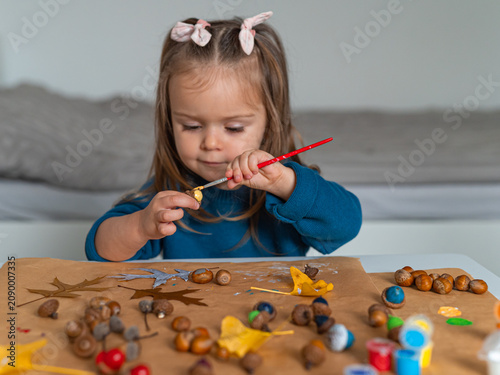 Cute toddler girl painting acorns with brush doing autumn DIY crafts
