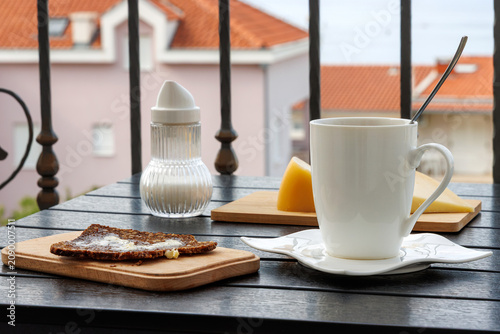 Balcony breakfast with coffee bread cheese and sugar dispenser. Mediterranean travel lifestyle scene with red rooftops in background

