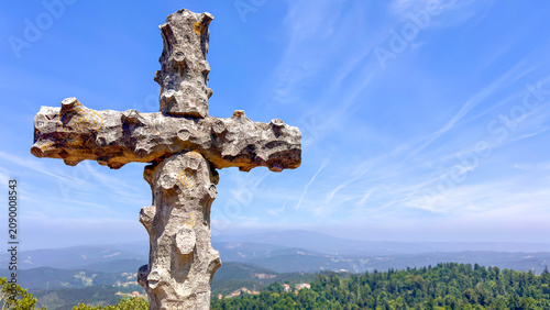Cross stands on a hilltop overlooking a landscape of trees and mountains under a blue sky, Portugal