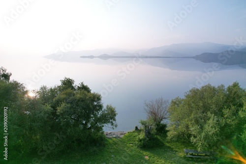 Fotografija Serene Lake View from Hilltop with Sunlit Trees and Distant Mountains