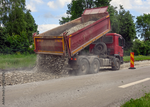 Red heavy dump truck unloading crushed stone or gravel onto a new road surface during construction works.