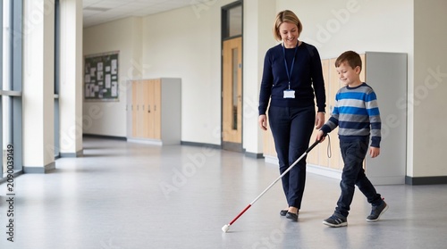 Teacher guiding visually impaired boy with white cane in school hallway  