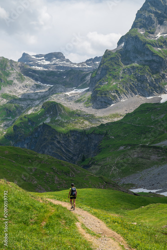 Obraz na plátně Lone backpacker walking a mountain path through green alpine meadows below rocky peaks and lingering snowfields near Engelberg, Switzerland