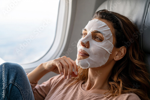 Young woman sits in an airplane window seat wearing a sheet face mask and looking out. Themes: travel wellness, self-care, in-flight skincare, relaxation, beauty routine, solo travel, mindfulness.