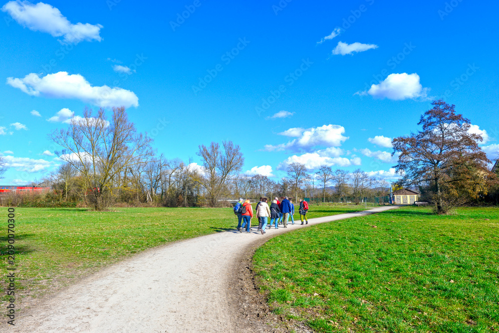 Obraz premium Gruppe von Wanderern mit Rucksäcken auf einem Spazierweg an einer großen Wiese im Frühling in Kahl am Main, Bayern