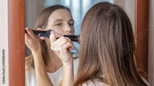 Young woman makes on her face using makeup brush while at home in a room looking at the mirror. Caucasian girl does makeup on her own.