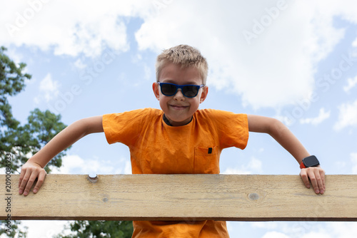 Tableau sur toile A boy in sunglasses looks down from above and holds onto a board with his hands against a background of a sky with clouds