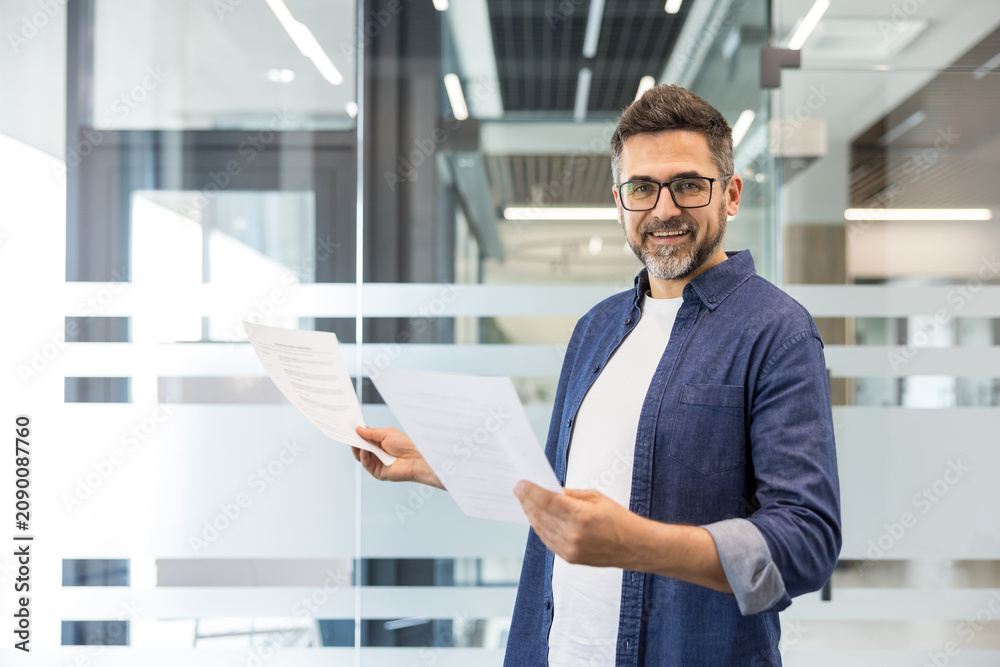 Fototapeta premium Confident businessman standing in a contemporary office corridor, holding various paperwork and smiling directly at the viewer, representing corporate success and professional communication