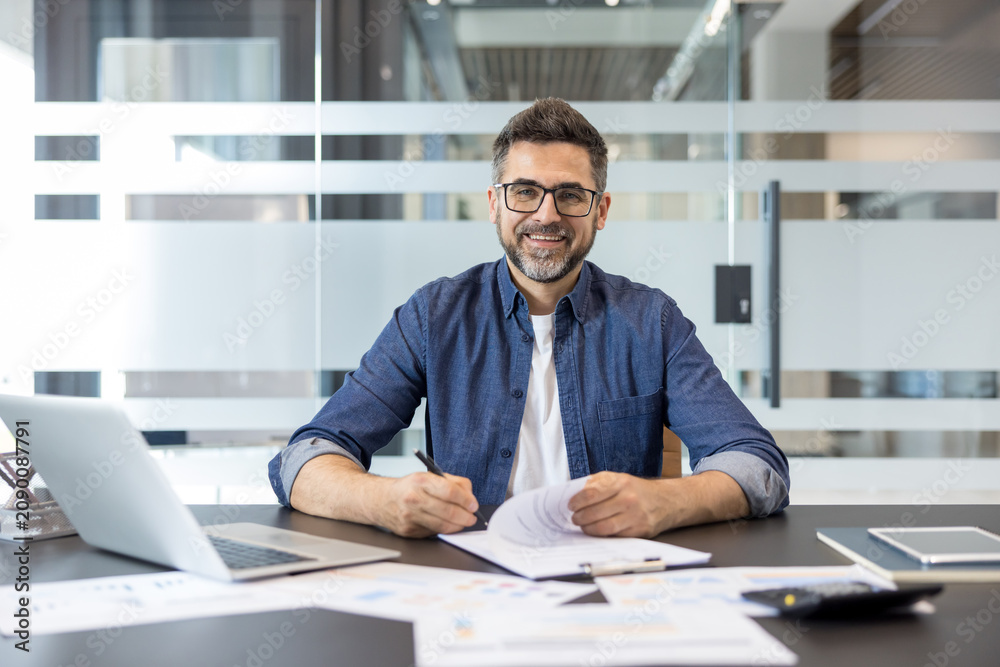 Obraz premium Confident businessman analyzing financial reports and signing paperwork, sitting at his desk with a laptop in a modern office, representing successful career and management