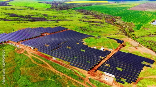 Aerial view of solar panel installation site. Dark photovoltaic modules bordered by green farmland and dirt path.