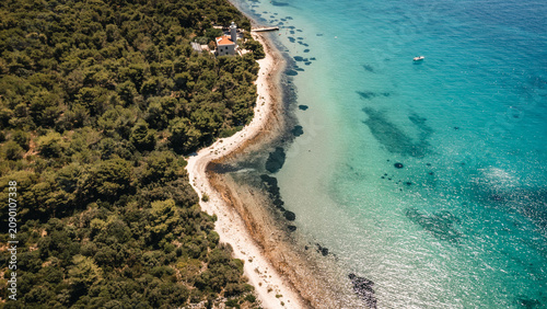 Aerial view rocky coastline with turquoise sea, dense pine forest and sunlit shore creating vivid contrast between clear water, sand, rocks and coastal greenery
