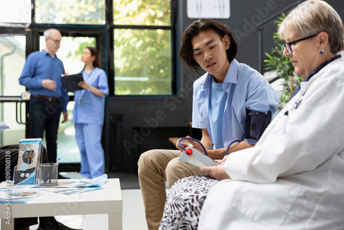 Konstfotografi Asian man wears arm blood pressure cuff in hospital lobby while female doctor analyzes vital signs on monitoring device