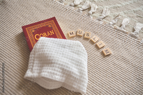 Quran, a white kufi, and scrabble letters spelling muslim on a beige prayer rug