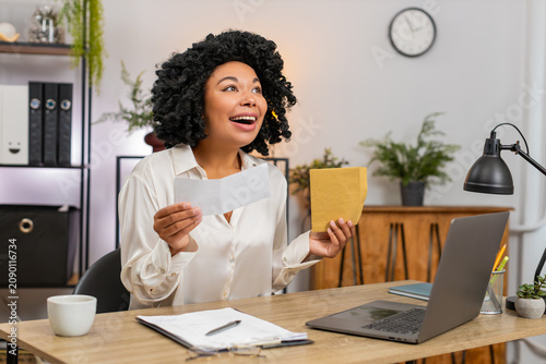 African American businesswoman at home office opens envelope reading letter then smiles wide celebrating job approval and big pay raise. Freelancer girl at table claps joyful excited for career step.