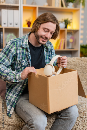 Slika na platnu Young man on home sofa opens box finds wireless headphones and checks new purchase with keen smile
