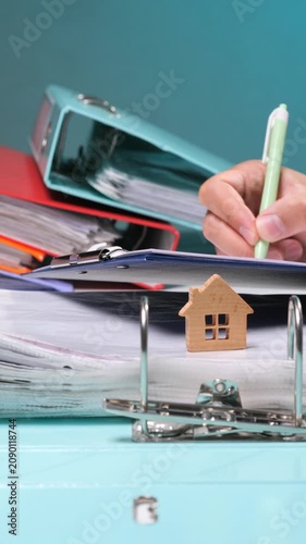 Vertical video hand writing on a clipboard with paperwork, a wooden house model resting on file folders, discussing home buying, mortgage, insurance, or property investment concepts