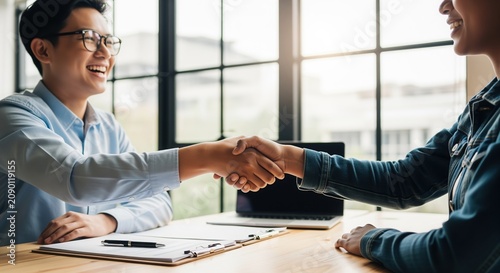 Two business professionals shaking hands across a desk in a bright office. One wears glasses and light blue shirt, the other a dark jacket. Ideal for depicting deals and partnerships.