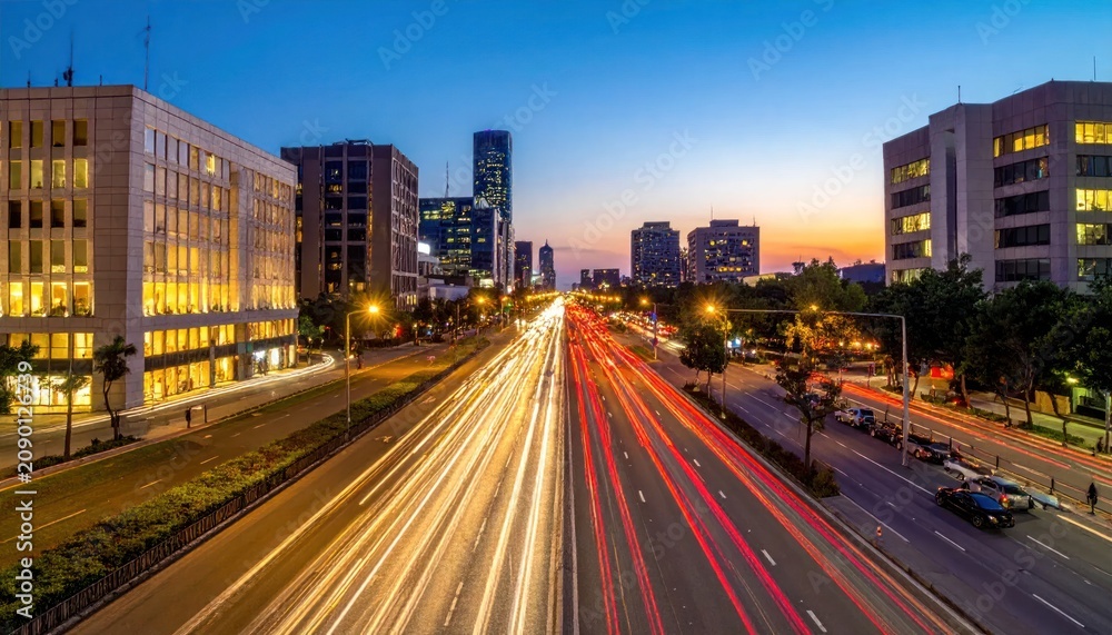 Obraz premium Cityscape at Dusk with Light Trails from Traffic on a Highway Lined with Modern Buildings and Trees Under a Blue Sky
