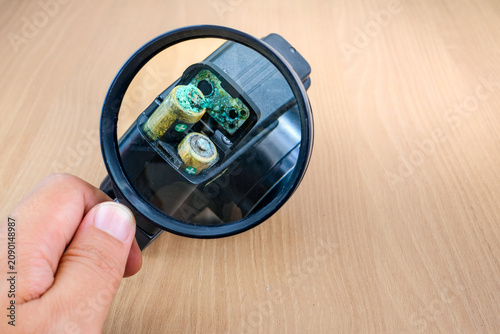A magnifying glass is used to inspect corroded batteries inside an electronic device on a wooden table. Suitable for battery leakage, electronic damage, corrosion inspection, device repair