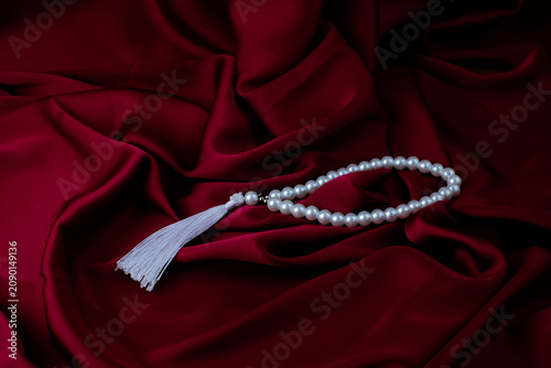 A white pearl prayer bead strand with a tassel is placed on dark red satin fabric. Suitable for Ramadan background, Islamic accessory, luxury fabric, spiritual still life